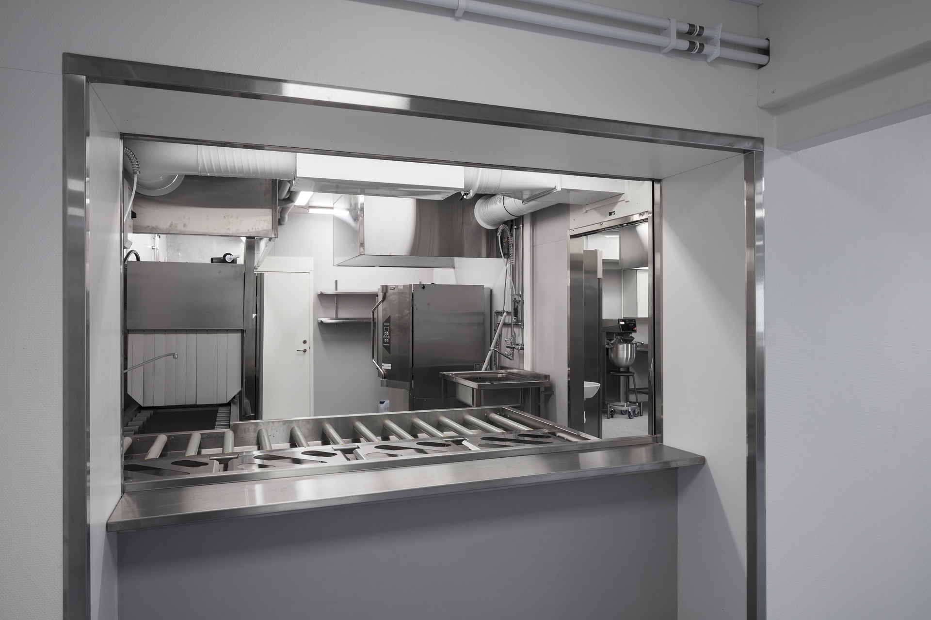 Serverings- och köksmiljö i skolans modullösning från Expandia, med rostfri serveringslucka, rullbana för brickor samt professionell köksutrustning anpassad för skolverksamhet.