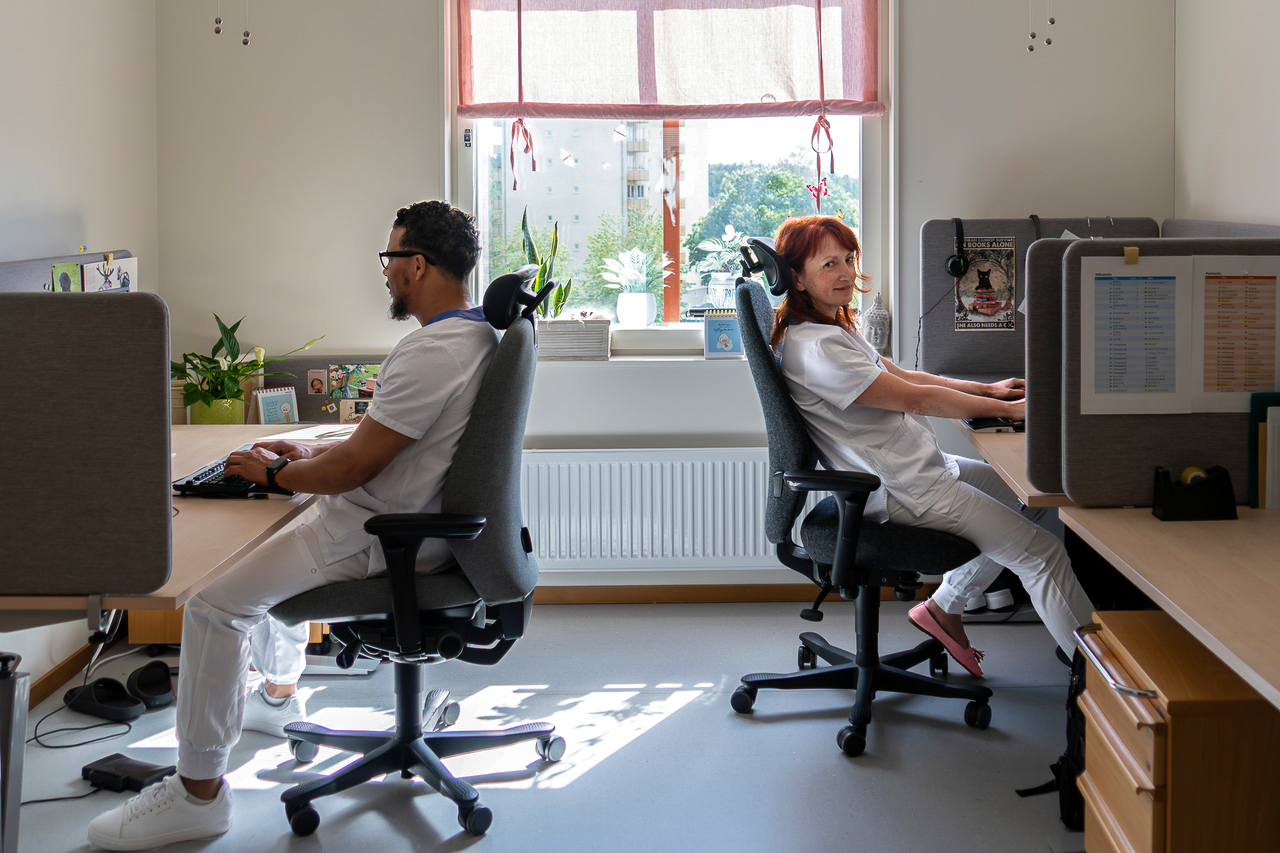 Två medarbetare arbetar vid separata skrivbord i kontorsmoduler från Expandia på Centralsjukhuset Karlstad. De sitter rygg mot rygg i ergonomiska kontorsstolar vid arbetsplatser med skärmar, personliga detaljer och gröna växter. Rummet är ljust med stort fönster som släpper in dagsljus och skapar en lugn arbetsmiljö.