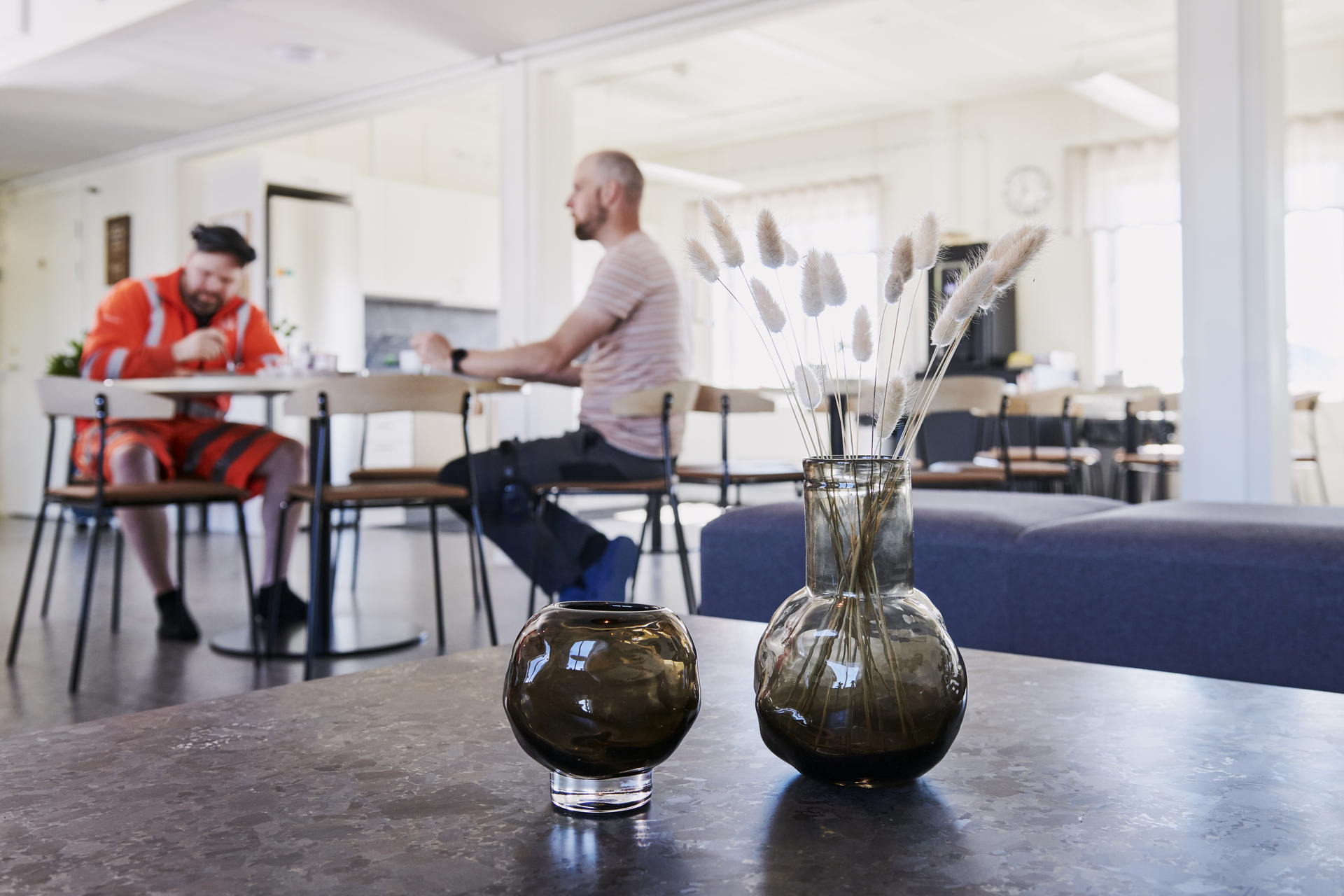 Pausutrymme i kontorsmoduler från Expandia med bord och stolar i en öppen och ljus miljö. I bakgrunden sitter två personer vid ett bord i samtal, medan en vas med torkade växter står i förgrunden. Bilden är tagen i kontorsmoduler vid Alviks trafik i Luleå.