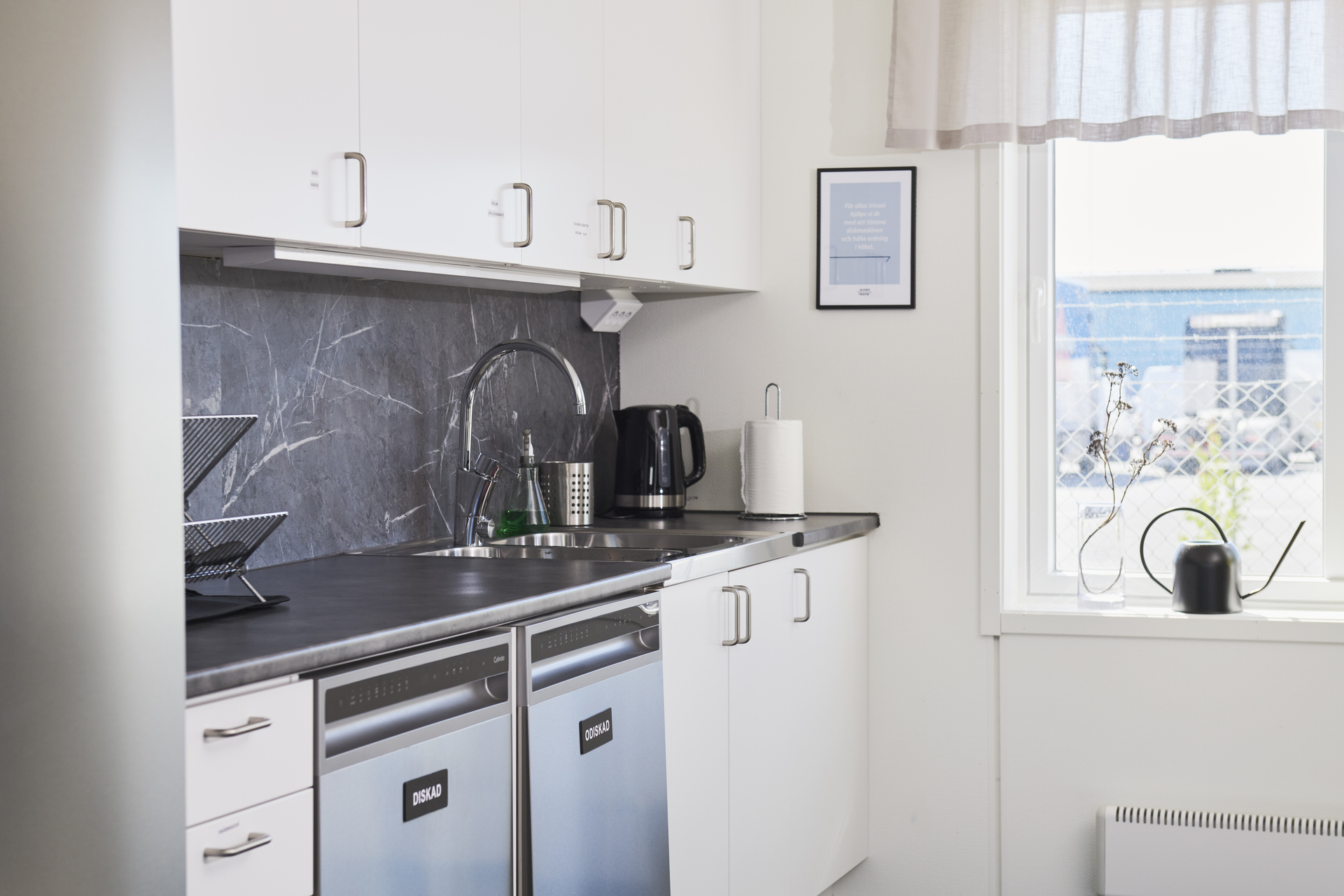 Kök i kontorsmoduler från Expandia med vita skåp, arbetsbänk, diskho och diskmaskiner. Rummet är ljust och funktionellt, med fönster som släpper in dagsljus. Bilden är tagen i kontorsmoduler vid Alviks trafik i Luleå.