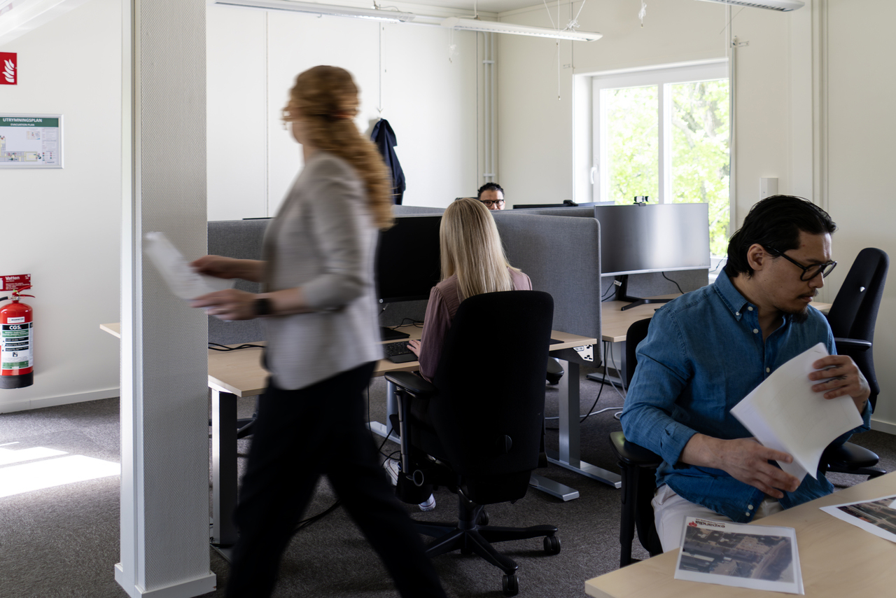 Kontorslandskap i kontorsmoduler från Expandia i Skanskas projektkontor i Karlstad. Flera personer arbetar vid skrivbord med skärmar, medan en person går förbi med dokument i handen. Rummet är ljust med fönster som släpper in dagsljus.