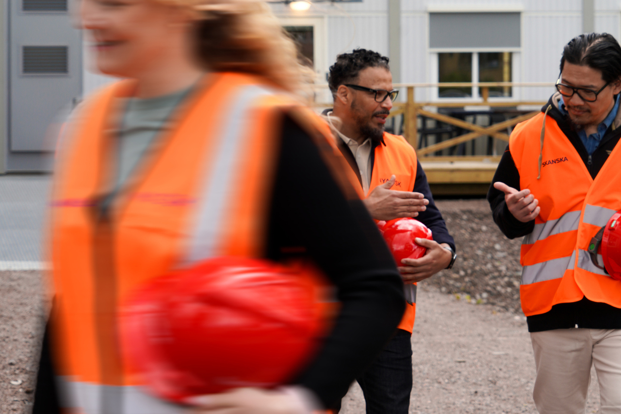 Personer i varselvästar och med skyddshjälmar i handen på väg ut från kontorsmoduler från Expandia vid Skanskas projektkontor i Karlstad. De samtalar medan de rör sig utomhus mot arbetsområdet, med modulerna i bakgrunden.