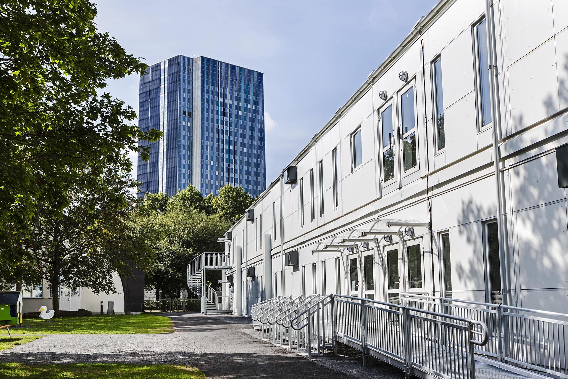 Skolmoduler från Expandia vid Sveaplansskolan i Stockholm. Ljus tvåplans modulrad med entréer, ramper och spiraltrappa, placerad i grön miljö med träd och gräsyta. I bakgrunden syns ett högt stadshus som kontrasterar mot skolområdet.