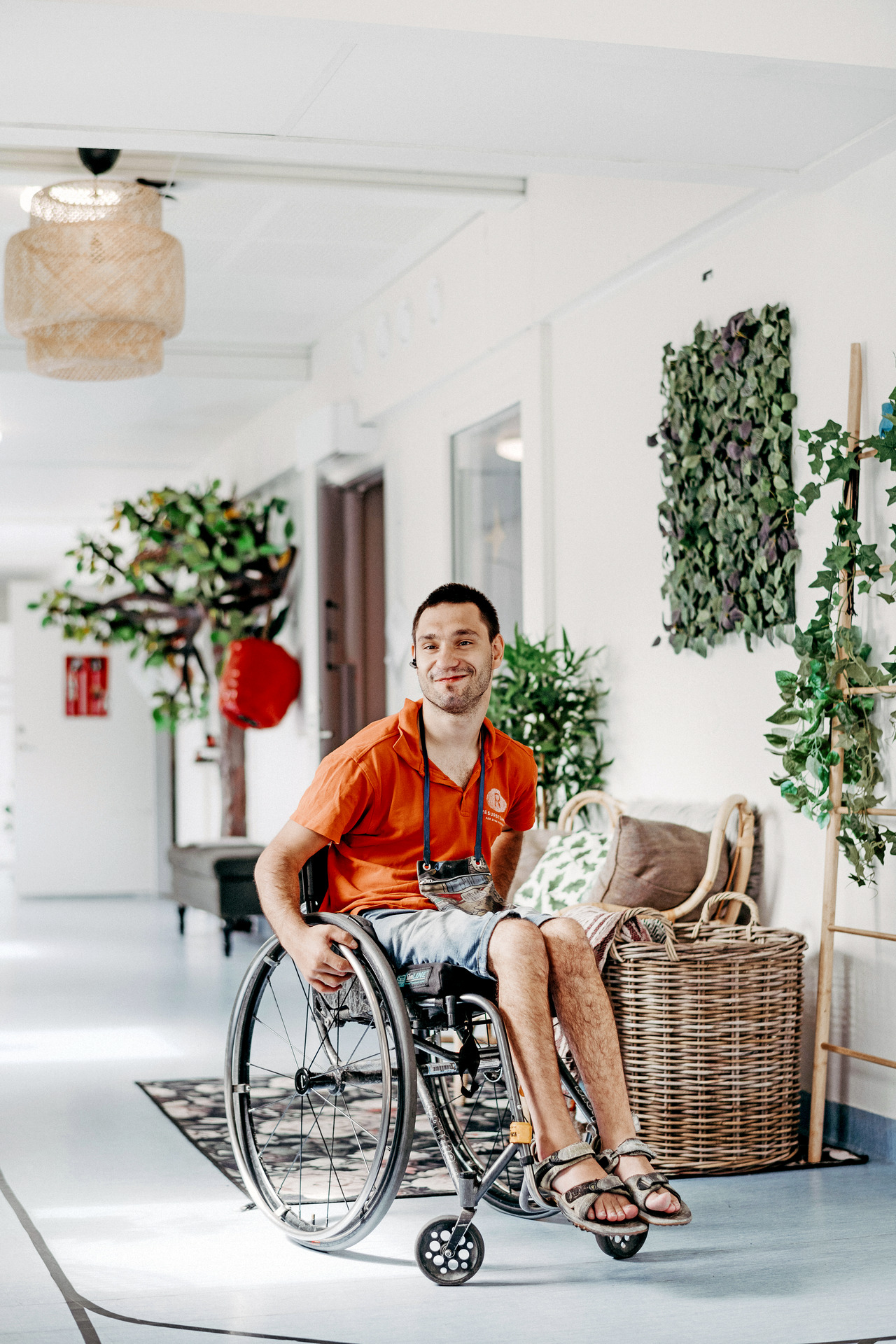 En person som sitter i rullstol ler i en ljus korridor med växter och sittmöbler vid Insperia daglig LSS-verksamhet i Ekerö.
