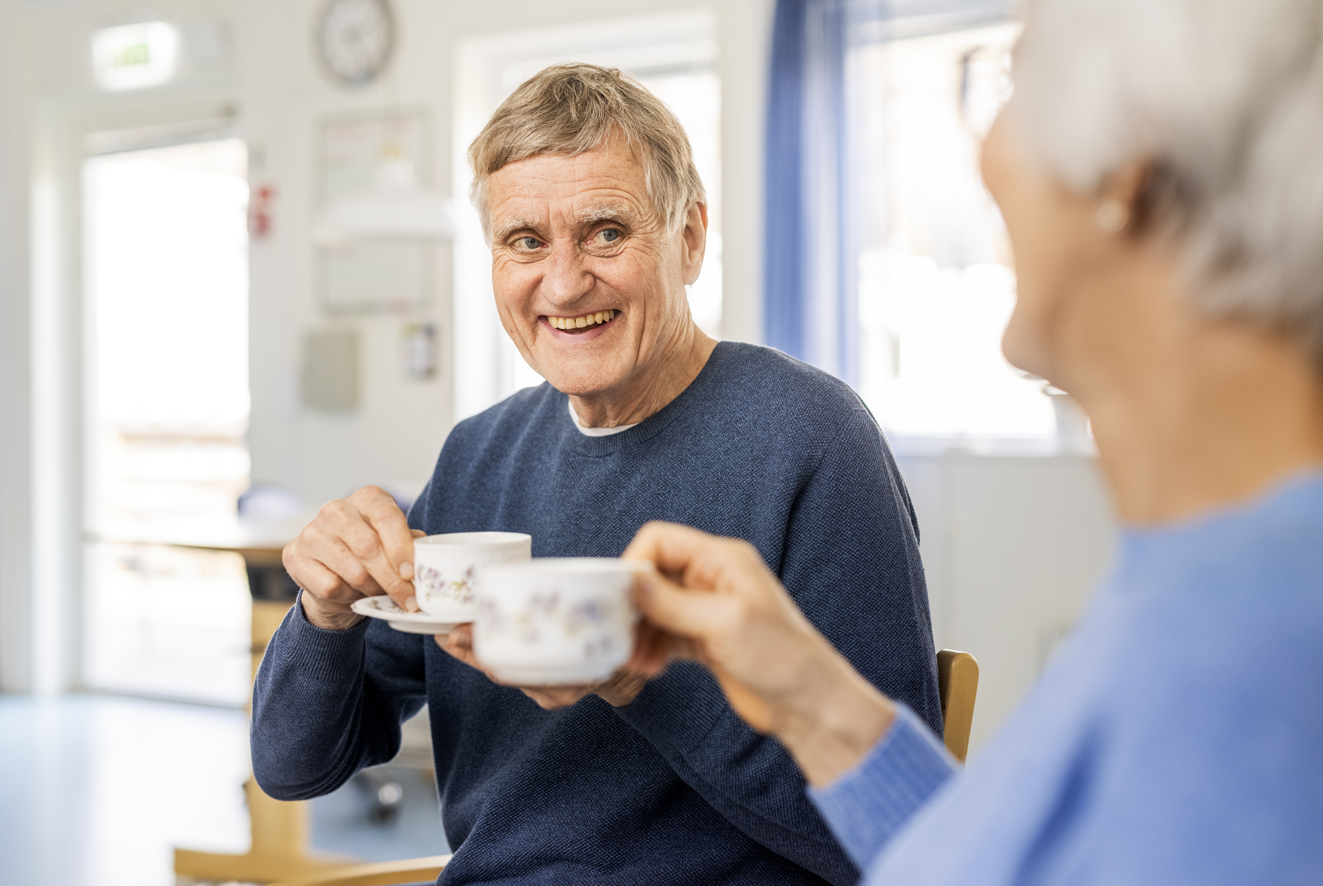 Två boende vid Borggården demensvård i Olofström sitter inomhus och dricker kaffe tillsammans. I bakgrunden syns en ljus och hemlik miljö som ingår i en modullösning från Expandia.