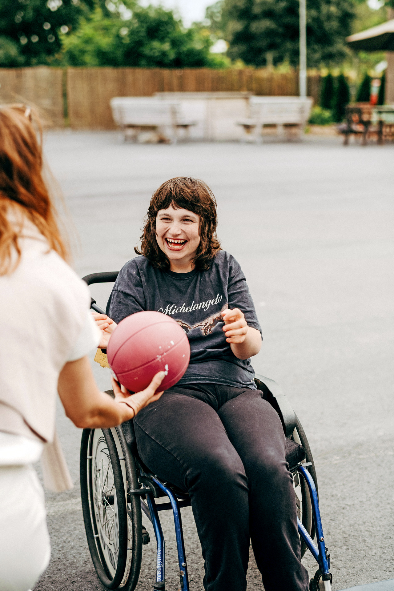 En person i rullstol skrattar och sträcker sig efter en röd boll som räcks fram av en annan person under en gemensam aktivitet utomhus.