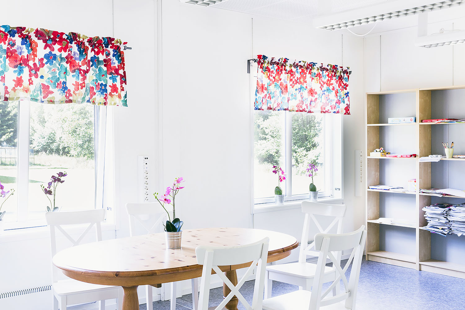 Ljust allrum i boendemoduler från Expandia med runt matbord och vita stolar, blomsterdekorerade gardiner vid fönstren samt öppen förvaring med hyllor längs väggen.