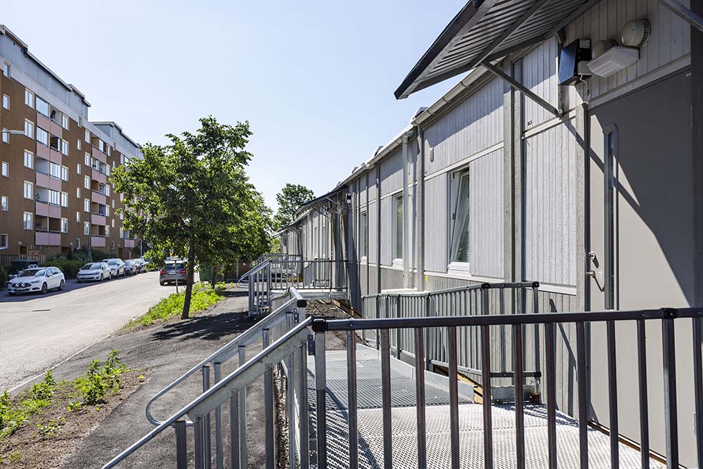Evakueringsboende i Henriksdal med modulbostäder längs gata, tillgängliga entréer med ramper och räcken samt flerbostadshus och parkerade bilar i bakgrunden.